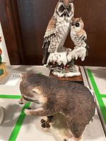 Two ceramic statues: an owl pair on a snowy branch with red berries and a raccoon standing on a base.