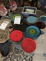 Multiple sets of metal coasters in various colors and designs, with small copper pot and a storage tin visible on a glass table.