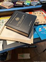 Photo showing closed black leather album titled 'U.S. First Day Covers & Special Covers' by Irene Muenchrath and other stamp materials on a table.