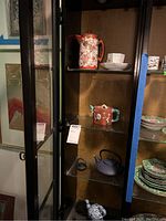 Glass cabinet showing shelves with multiple Asian style teapots, porcelain plates, and a white cup and saucer set.