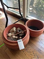Two large round terracotta planters on hardwood floor near a window, one filled with soil, rocks, and a stone artifact, the other empty.