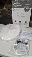 Photo showing unopened white American Standard AquaWash elongated bidet seat in original packaging with box and parts on white surface.