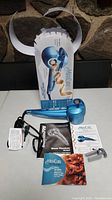 Photo showing blue BaBylissPRO Nano Titanium MiraCurl curling machine with power cord, original box, and three instruction manuals along with small brush and clip accessory.