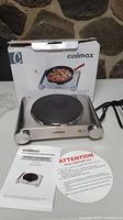 Front view of Cuisimax stainless steel hot plate next to box, with power cord, manual, and safety instructions.