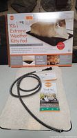 Photo showing the K&H Extreme Weather Kitty Pad box with picture of a cat, the heated pad, steel wrapped power cord, and included fleece cover.