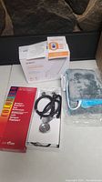 Photo showing two boxed items: white box with heating pad packaging and red box labeled Sprague-Rappaport stethoscope. A blue heating pad is visible outside one box along with the stethoscope tubing and chestpiece.