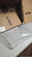 The two Kraus branded cardboard boxes for sink grids visible, one partially open with the metal sink grid visible inside clear plastic wrapping on a countertop.
