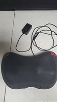 Top down view of black TheramMedic shiatsu neck massager pillow with power adapter next to it on white surface.