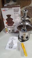 Chocolate fountain assembled with three stainless steel tiers on a stone background, box and accessories placed beside.