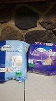 Two opened packages of TENA incontinence briefs, one blue labeled Bariatric Ultra and one purple labeled Over Night, placed side by side.