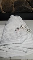 Folded white quilted heated mattress pad topper with attached controller and cables laid on white surface in front of a stone wall background.
