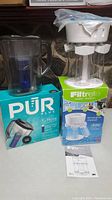 Photo showing the PUR Plus water jug next to its box and the Filtrete water filtration system with its components on top of its box. Also includes a manual.