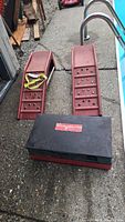 Pair of red painted steel automotive ramps with textured footholds and safety handles positioned on ground next to the black parts washer.