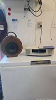 Photo showing the Bose Wave Music System with digital display clock, standing on a surface beside a round wooden portable speaker with handle and black speaker cone.
