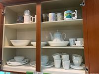 Photo showing upper shelves with miscellaneous mugs including ceramic and patterned mugs, and white teapot with matching small dishes.