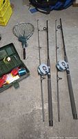 Overall lot showing two fishing rods with attached Daiwa reels, green fish net, and open tackle box with various fishing items visible.