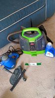 Front view of Bissell Little Green ProHeat Pet cleaner, blue cleaning solution bottle, Omega handheld steamer and several attachments on carpet near blue furniture.
