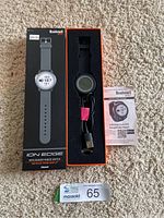 Full view of the Bushnell Ion Edge GPS rangefinder watch in box showing watch, USB charging cable, packaging, and quick start guide on carpeted floor.