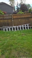 Three aluminum ladders resting on grass including a 14-foot extension ladder lying flat and a 9-foot extension ladder leaning against a fence with a 10-foot folding ladder partially visible.