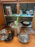 Wide view of cookware including stockpot, egg poacher pan, black non-stick frying pans, metal colanders, steamer insert and pot watcher disc.