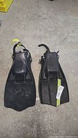 Two black scuba diving fins side by side on floor, one pair visibly Scubapro and one Aqua-Lung