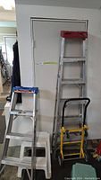 Photo showing the 6-foot aluminum step ladder, 4-foot aluminum step ladder, yellow and black hand truck, and two small step stools in the corner of a room.