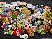 Top view of an assortment of retro pinback buttons showing a mix of slogans, logos, and cartoon character designs scattered on a black surface.