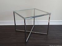 Photo of entire side table on dark wood flooring showing the chrome frame and clear glass top from a slight angle.
