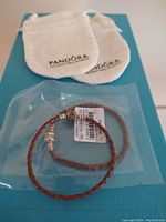 Two braided leather Pandora charm bracelets shown on a blue surface with two white Pandora branded drawstring pouches behind. One bracelet is sealed in plastic packaging with a tag and barcode visible.