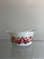 Front view of white Pyrex casserole dish decorated with red and orange floral and bird pattern with clear glass lid.