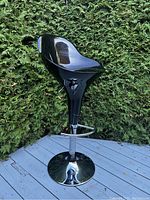 Side angled view of black glossy curved seat with adjustable height lever, chrome pedestal, footrest, and round base outdoors with hedge background.