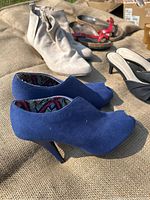 Photo showing multiple pairs of women's shoes including beige suede booties, blue suede peep-toe booties, black open-toe mules, and sandals in tan and brown with red straps.