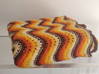 Folded vintage hand knitted blanket showing multi-color wavy zigzag pattern in shades of orange, yellow, brown, and white.