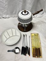 Full display of all fondue set components including pot, burner stand, ceramic dish, skewers, and measuring spoon