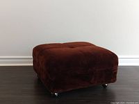 Front left angle view of the brown plush ottoman showing the tufted top and caster wheels.