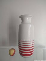 Full view of tall white ceramic vase with flared mouth and horizontal red stripes, shown next to an apple for scale.