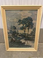 Framed textile art depicting a landscape with trees, a river, and houses under a cloudy sky. Mounted in a cream wooden frame.