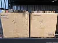 Two cardboard boxes labeled "1" and "2" filled with vinyl records standing upright on a metal shelf.