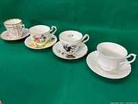 Four distinct tea cups and their saucers arranged in a row on a green surface, showing differences in colors and floral patterns.