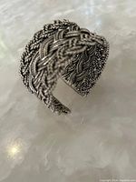 Close-up photo of the silver tone bracelet showing detailed braided wire design on a light marble-like surface.