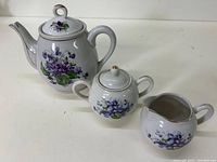Full view of ceramic teapot, sugar bowl with lid, and creamer all decorated with violets and gold trim.