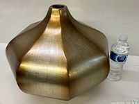 Full view of a large polygonal brass vase with a narrow top opening beside a water bottle for scale, showing the brushed texture and overall shape.