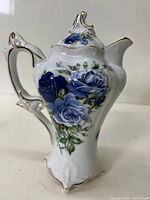 Front view of a tall white porcelain coffee pot featuring blue rose floral decoration and gold trim accents on spout, lid, and handle.