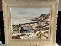 Full view of the framed oil on board painting depicting a rustic barn near the ocean with cliffs in the background.