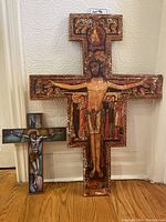 Photo showing both religious crosses side by side against a wall and hardwood floor, including a large wooden painted cross with Christ and figures, and a smaller stained glass style cross.