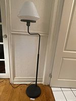 Full view of the black metal floor lamp standing against a wall showing its base and adjustable arm with white glass shade.