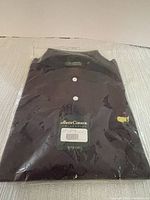 Black golf shirt in original plastic packaging, neatly folded, showing Amen Corner Collection tag with price sticker $79