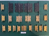 Overview of all 21 assorted metal and brass hinges laid out on green fabric. Includes three sizes with brass and black finishes.