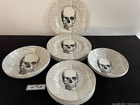 View of 14 pottery plates featuring black skull illustration on handwritten script background, arranged in groups.