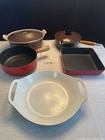 All six cookware pieces on table: two light grey Le Creuset pots with lids, four Staub pans in red and brown.